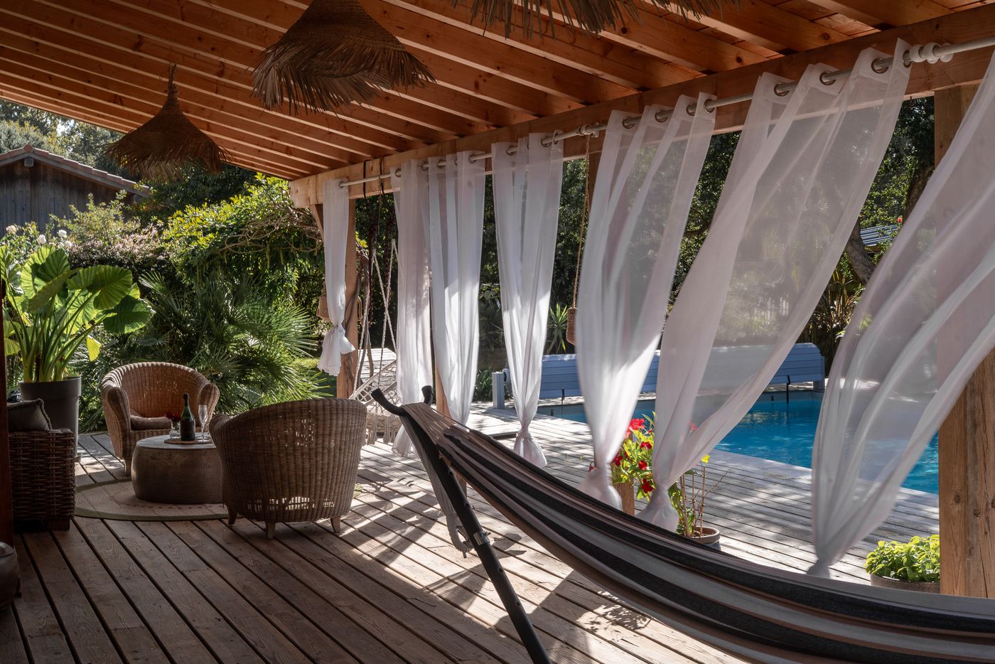 Salon sous la pergola avec fauteuils en rotin et hamac, piscine en arrière-plan